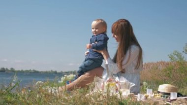 Anne-çocuk ilişkisi, neşeli bir kadın aile pikniği sırasında oğluyla oynuyor. Nehir kenarındaki çayır çimlerine yakın bir yerde.