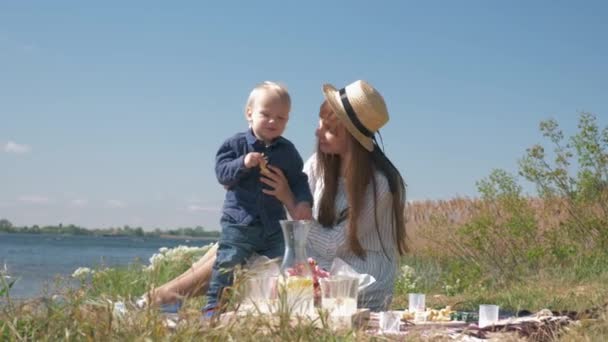 Glad barndom, rolig pojke äter citron och leker med ung mamma under familjen picknick nära floden mot himlen — Stockvideo