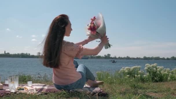 Щаслива довгошерста жінка насолоджується красивим букетом на природі під час пікніка на газоні річкою в сонячну погоду — стокове відео