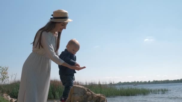 快乐的年轻母亲戴着草帽，带着蹒跚学步的男孩，一边在河边休息一边扔小鹅卵石，一边享受娱乐 — 图库视频影像