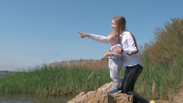 Joven feliz hembra con niño mirar a la distancia y mostrar la mano en la distancia lago de pie sobre piedra en el agua — Vídeo de stock