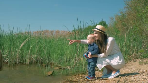 Młody szczęśliwy mama pokazuje natura dla dziecko chłopiec podczas odpoczynku na brzegu jeziora z trzciny i patrząc w oddali — Wideo stockowe