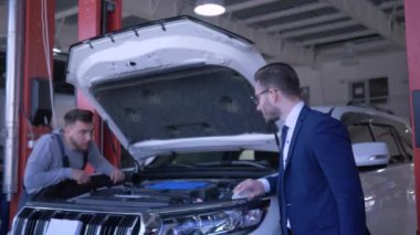 Profesyonel araba bakımı, teknisyen açık kaputlu aracı tamir ediyor ve araç sahibi benzin istasyonunda olumlu bir hareket gösteriyor.