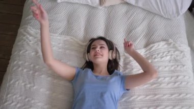 Neşeli bir kadın kulaklığı takıp yatağa düşüyor ve evde dinlenirken cep telefonundan ve keyif müziğinden şarkılar dinliyor.