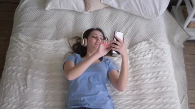 Neşeli kadın kulaklıkları takıyor müziğin tadını çıkarıyor ve yatakta dinlenirken cep telefonuyla selfie çekiyor.
