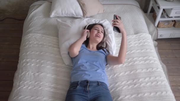 Χαρούμενο κορίτσι σε ακουστικά με κινητό τηλέφωνο απολαμβάνουν μουσική ενώ ξεκουράζονται στο κρεβάτι στο σπίτι για διακοπές, top view — Αρχείο Βίντεο