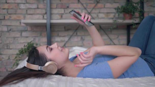 Bella donna in auricolare con telefono cellulare deliziare la musica mentre si riposa sul letto a casa in vacanza — Video Stock