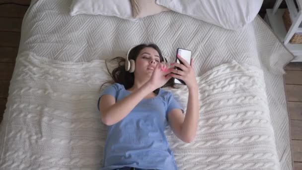 陽気な女性がヘッドフォンで音楽を楽しみ、ベッドでリラックスしながら携帯電話で自撮り写真を撮る — ストック動画