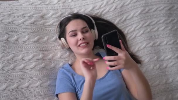 Agradável sorrindo fêmea em fones de ouvido com telefone ter música prazer e canta enquanto relaxa na cama em casa closeup no fim de semana, vista superior — Vídeo de Stock