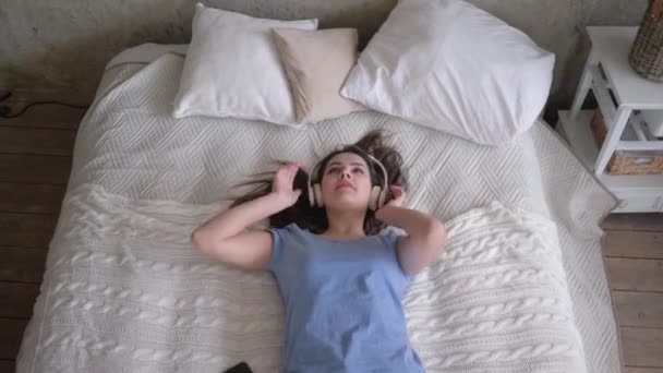 Χαλαρώστε το χρόνο, χαρούμενη κοπέλα σε ακουστικά πέφτει στο κρεβάτι και ακούει τραγούδια από το smartphone και απολαμβάνοντας τη μουσική, ενώ ξεκουράζεται στο σπίτι — Αρχείο Βίντεο
