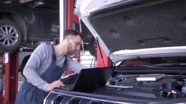 Computer diagnostics of cars, young mechanic male uses laptop technology while repairing vehicle with open hood at service station — ストック動画