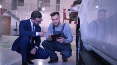 Tabletli teknisyen erkek, oto tamirhanesinde araba tekeri yakınında tüketiciyle iletişim kuruyor.