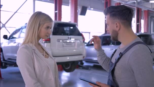 Reparateur en klant vrouw communiceert over auto reparaties op de achtergrond van rijzende auto op lift bij tankstation — Stockvideo