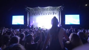 Kalabalık taraftarları el sallıyor ve gece büyük ekranlı parlak sahnelere karşı rock canlı müzik gecesinde akıllı telefondan fotoğraf çekiyor.