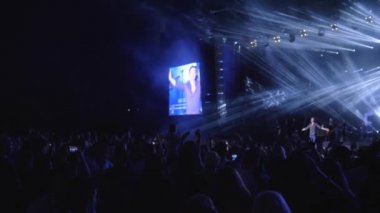 Hayranlar, gece rock konseri sırasında sahne ışıkları eşliğinde parlak sahnede müzisyenlerle el çırpıyor.