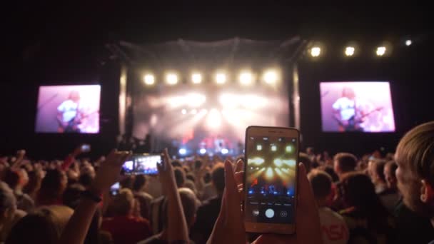 팬들은 휴대폰으로 사진 과 비디오를 찍는다. 록 콘서트는 어두운 밤에 대형 스크린을 갖춘 밝은 무대를 배경으로 한다. — 비디오