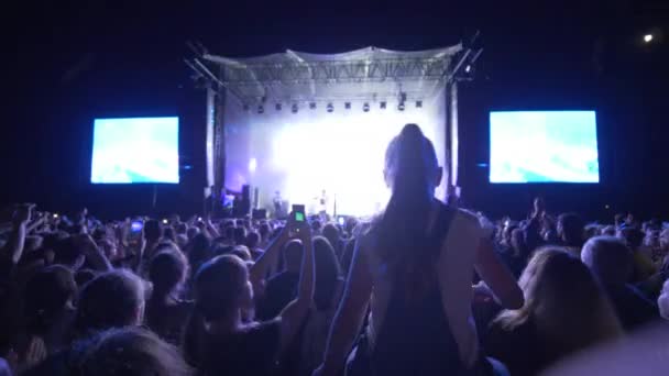 Torcedores de multidão acenam as mãos e fazem fotos no smartphone na noite de música ao vivo contra o palco iluminado com telas grandes à noite — Vídeo de Stock