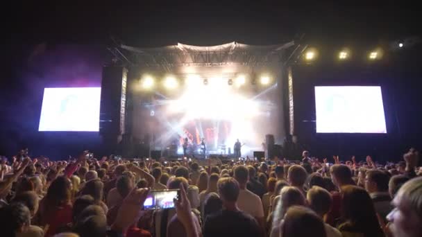 Толпа людей на концерте, много молодежи делают фотографии и видео на мобильный телефон на фестивале живой музыки рок против ярко освещенной сцене с большими экранами ночью в темноте — стоковое видео