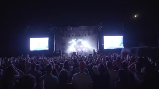 Crowd people jump and clap hands at rock live music concert against brightly lit stage with large screens at night — Stock Video