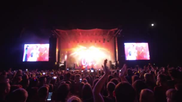 Публика отдыхает на рок-концерте против ярко освещенной сцены с большими экранами ночью — стоковое видео