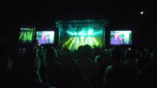 Ventilador con teléfono móvil en las manos hace video de concierto de rock en vivo entre la multitud contra la escena brillantemente iluminada con focos en la noche — Vídeos de Stock