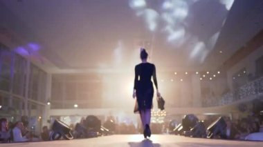 Moda sunumu, uzun bacaklı, topuklu ayakkabılı, siyah şeffaf elbiseli bir manken podyumda seyircilerin gözü önünde yürür.