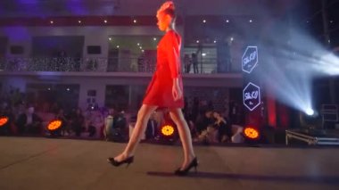 Moda şovu, model kız güzel kırmızı elbiseli yüksek topuklu ayakkabılı kız podyumda spot ışıklarına doğru yürüyor.