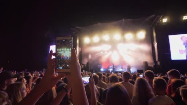 Вентилятор со смартфоном в руке записывает видео рок-фестиваля в ночное время в ярких огнях сцены на фоне толпы — стоковое видео