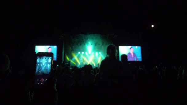 Ventilador com smartphone em mão grava vídeo sobre a multidão de fundo em luzes de palco brilhantes no festival de rock à noite — Vídeo de Stock