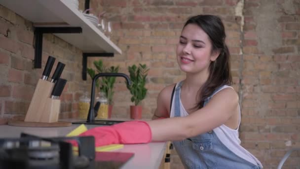 Feliz joven ama de casa en guantes de goma para limpiar los muebles polvorientos en la cocina — Vídeos de Stock