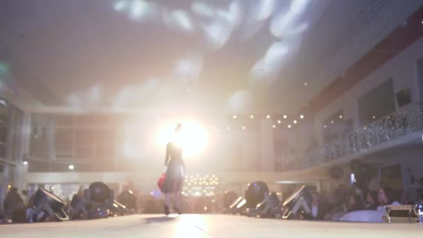 Desfile de moda, modelo podio mujer en zapatos de tacón alto en negro elegante vestido camina en pasarela en el centro de atención brillante en frente de la gente en desenfocado — Vídeos de Stock