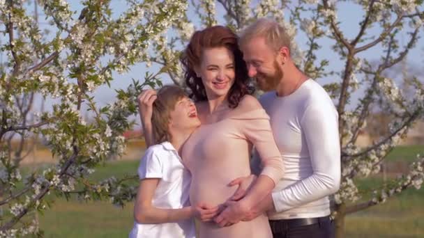 Feliz familia alemana, hombre sonriente con la mujer embarazada y el hijo adolescente abrazándose en floreciente jardín de primavera cerca del árbol frutal — Vídeo de stock