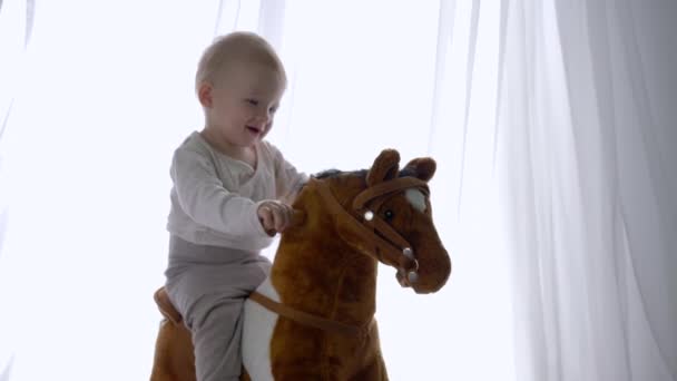 Счастливое детство, улыбающийся ребенок сидит и качается на игрушечной лошади дома — стоковое видео