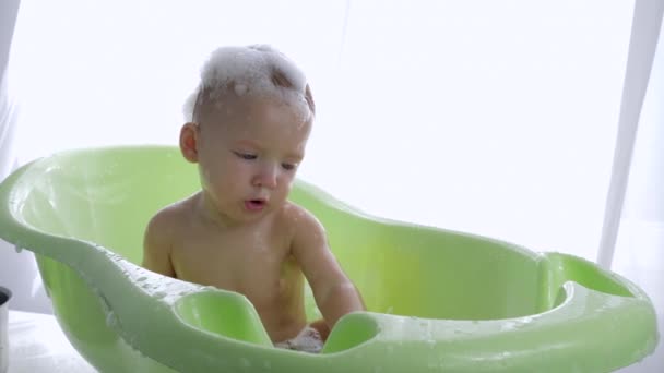 Procédures d'hygiène, curieux enfant en bas âge en bonne santé dans la mousse goûte assis dans l'eau dans le bain dans une pièce lumineuse — Video