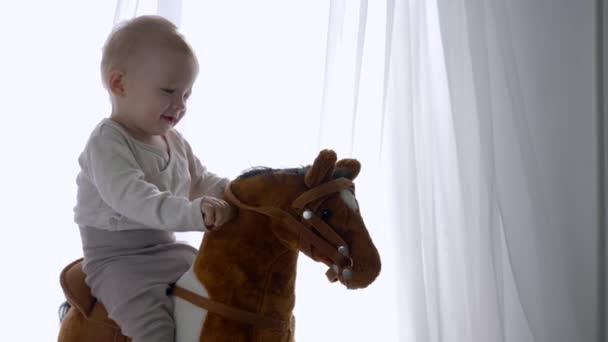 Раннее детство, счастливый мальчик, качающийся на игрушечной лошади дома — стоковое видео