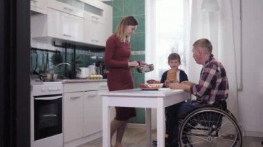 Aile yemeği, tekerlekli sandalyede mutlu bir baba, mutfakta gülümseyen bir eş ve oğluyla yemek hazırlamak.