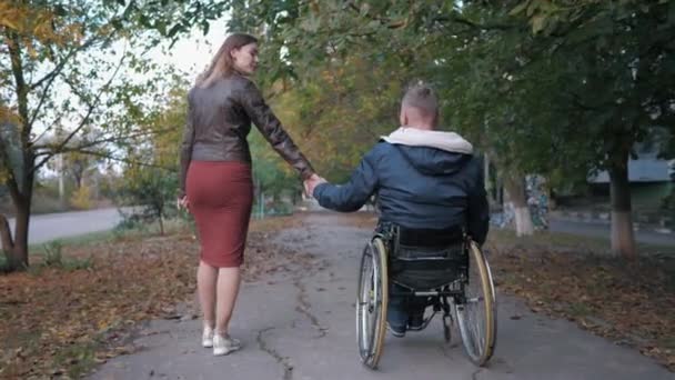 Amado discapacitado en silla de ruedas sostiene a sus esposas de la mano mientras camina en el parque de otoño entre los árboles — Vídeos de Stock