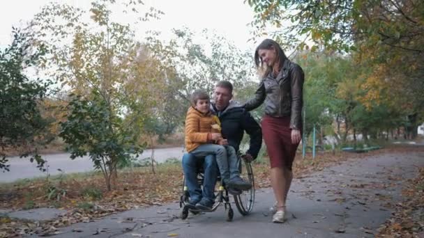 Disabled man in a wheelchair rides a boy on the cradle while walking along autumn street with his girlfriend — Stock Video