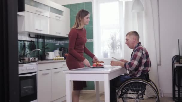 Family atmosphere, a husband in wheelchair cooks in the kitchen with his loving wife — Stock Video