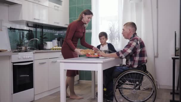 Interação familiar, um homem em uma cadeira de rodas em um lugar com sua esposa e filho preparar comida na cozinha, falar bem e sorrir — Vídeo de Stock