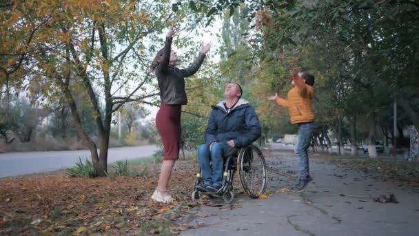 Сімейне дозвілля, усміхнений інвалідний чоловік в інвалідному візку з щасливою дружиною і дитиною викидає красиве осіннє листя на тлі дерев — стокове відео