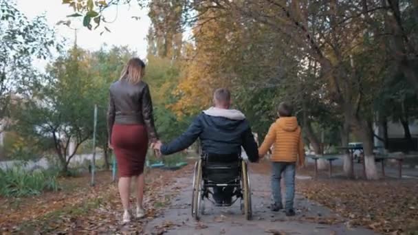 Familjerelationer, lycklig handikappad person i rullstol njuter av livet tillsammans med barn och hans fru sträcker upp händerna medan de går på vacker hösteftermiddag, bakifrån — Stockvideo