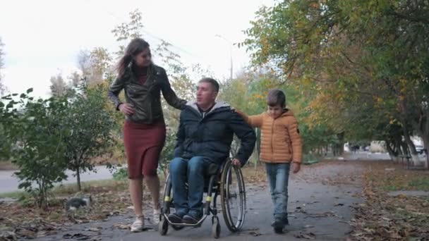 Rodinné vztahy, šťastný otec zdravotně postižené osoby na invalidním vozíku procházky se svou veselou rodinou podél podzimní ulice — Stock video