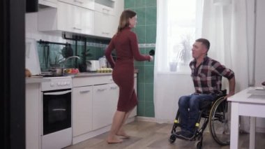 Tekerlekli sandalyedeki mutlu engelli adam mutfakta kız arkadaşıyla dans ederken eğleniyor.