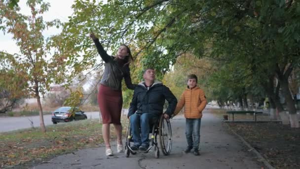 Sostegno familiare, marito disabile in sedia a rotelle cammina con la famiglia in una bella giornata autunnale per le strade della città — Video Stock