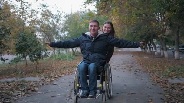 Apoyo amistoso, hombre discapacitado feliz divertirse en silla de ruedas paseo sonriendo hembra en el parque de otoño — Vídeos de Stock