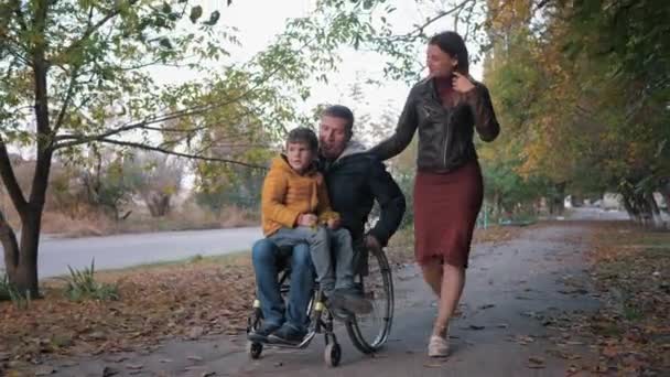 Щасливе батьківство, дитина сидить на колінах свого батька-інваліда в інвалідному візку під час прогулянки з матір'ю на тлі осінніх дерев — стокове відео