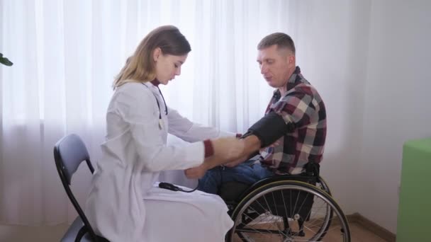 Hälso- och sjukvård, en patient i rullstol undersöker läkaren mäter trycket på honom med en medicinteknisk produkt — Stockvideo