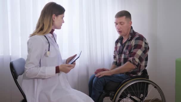 Чоловічий пацієнт у інвалідному візку під час медичного огляду розповідає про свої проблеми зі здоров'ям до лікаря-жінки — стокове відео