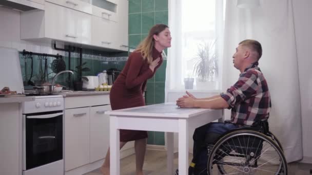 Нещасний інвалідний чоловік в інвалідному візку в стресовій ситуації виявляє відносини зі своєю нервовою дружиною, махаючи руками агресивно на фоні кухні в квартирі — стокове відео